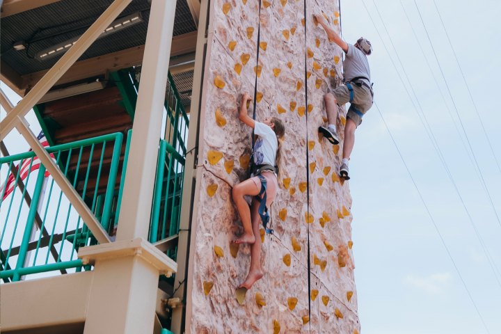 Two people rock climbing on an outdoor climbing wall with safety harnesses.