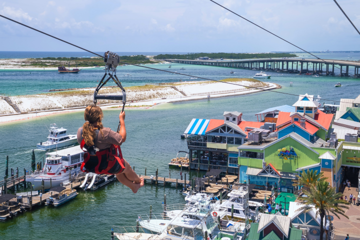 Person ziplining over a marina with colorful buildings and boats, near a sandy beach and bridge.