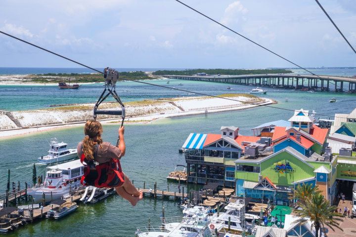 Person ziplining over a marina with colorful buildings and boats, near a sandy beach and bridge.