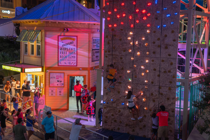 People climbing an illuminated rock wall near a ticket booth at night.