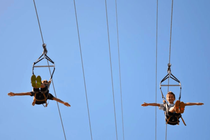 Two people ziplining in harnesses with arms outstretched, against a clear blue sky.