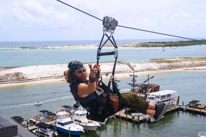 Person ziplining above water and docks, wearing a bandana, sunglasses, and casual attire.